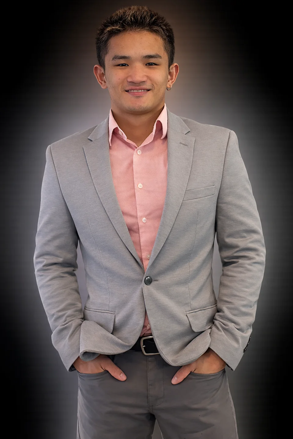 Portrait in a gray suit and pink shirt against a dark studio-style background.